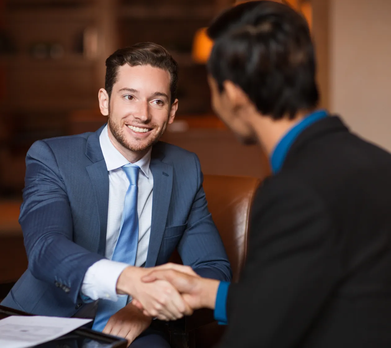 Two businessmen shaking hands during a meeting, one smiling and wearing a blue suit and tie.