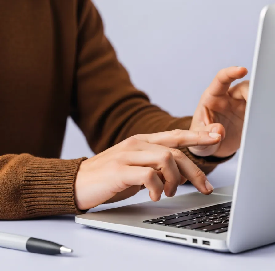 Person typing on a silver laptop keyboard wearing a brown sweater with a pen lying on the table.