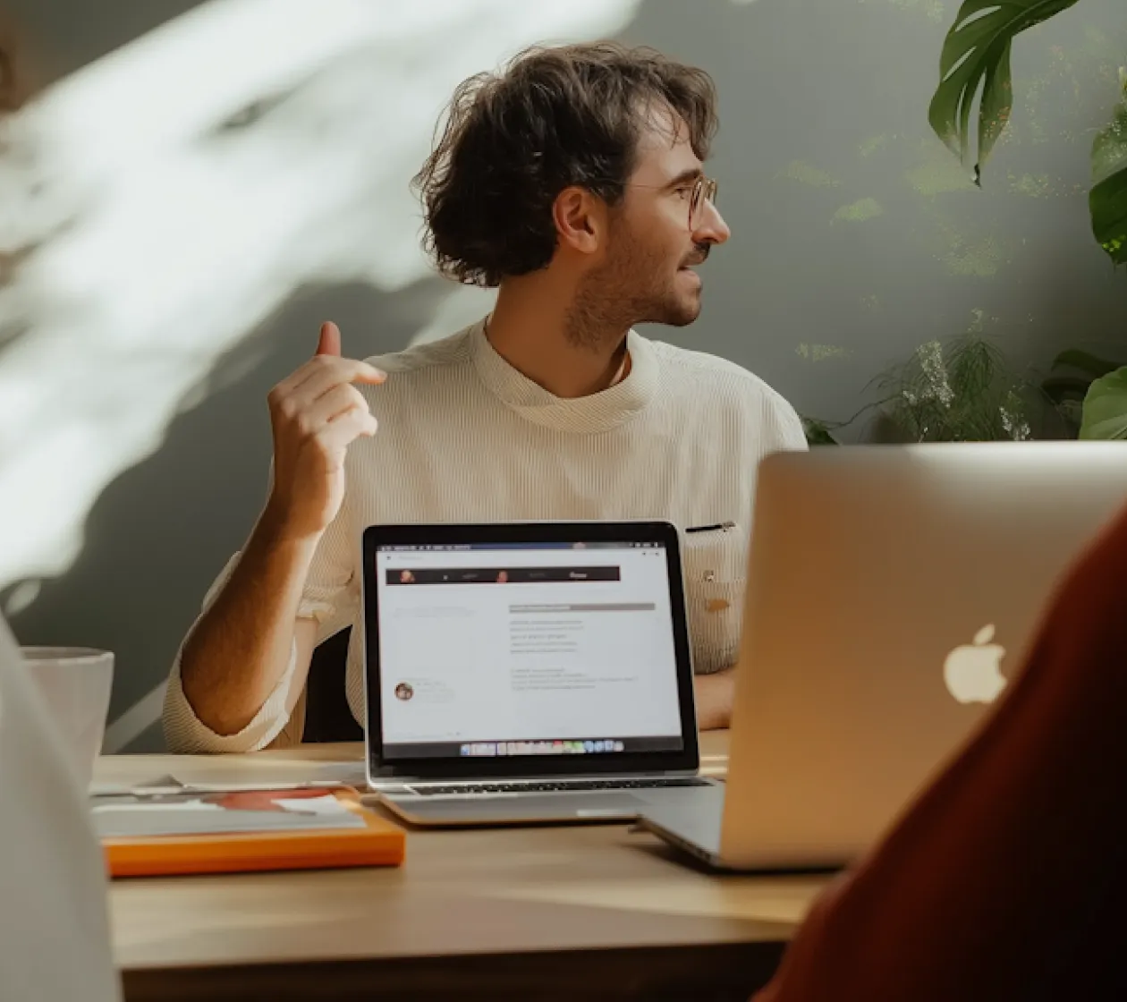 Man wearing glasses and a light sweater seated at a table with two laptops, gesturing with his hand.