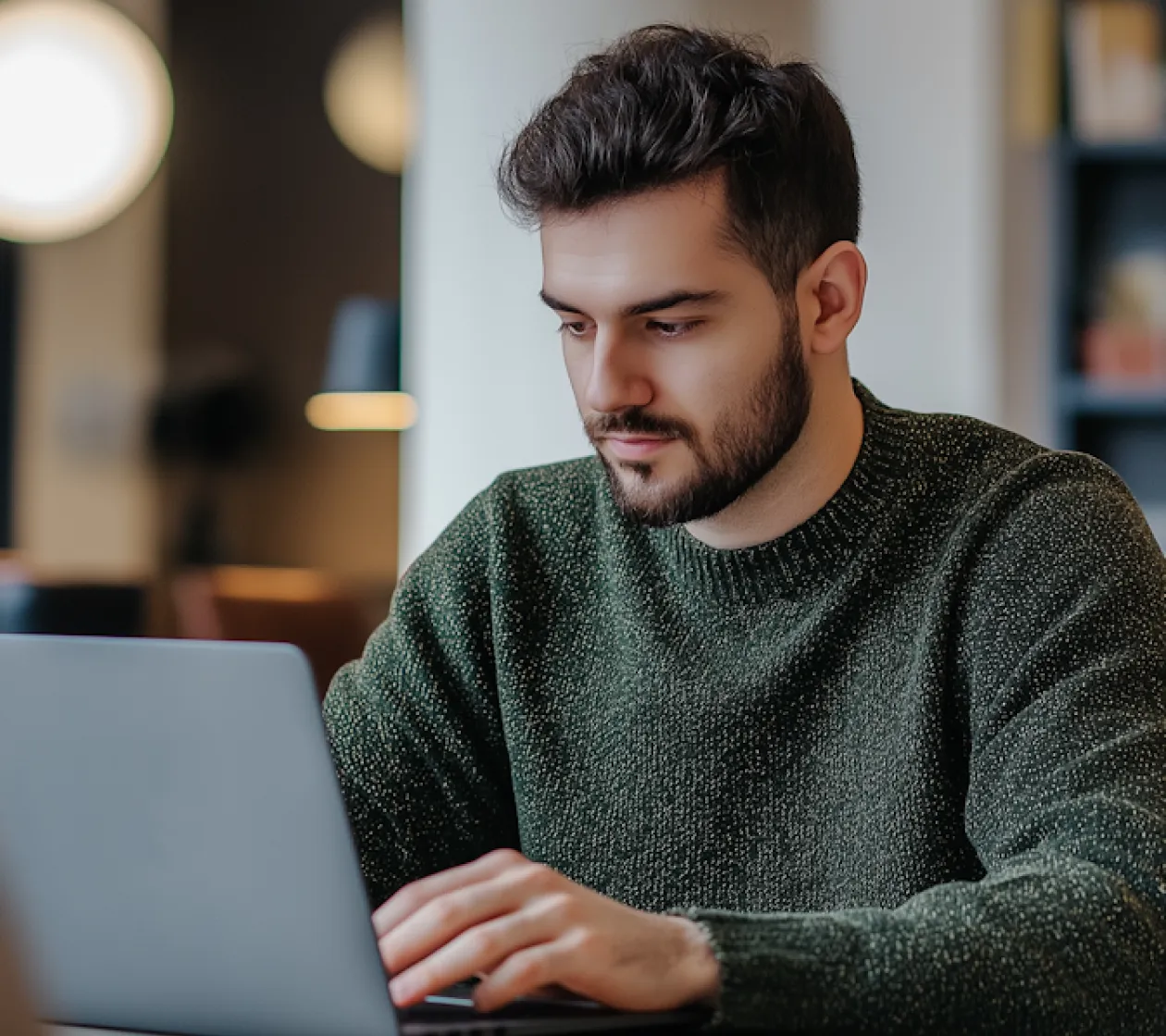 Bearded man wearing a green sweater working on a laptop in a cozy indoor setting.