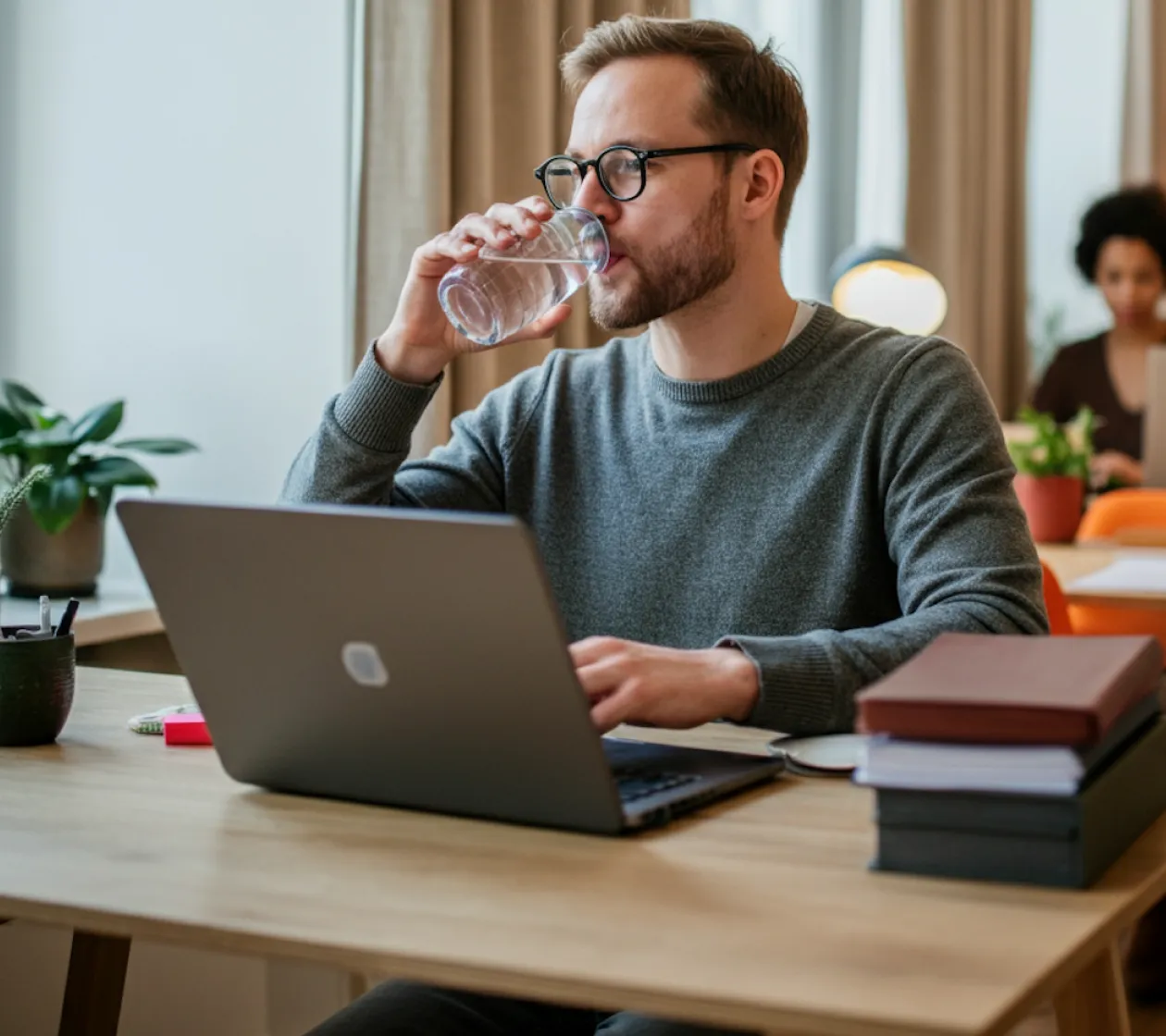Man with glasses drinking water while working on a laptop at a desk with stacked books and a plant.