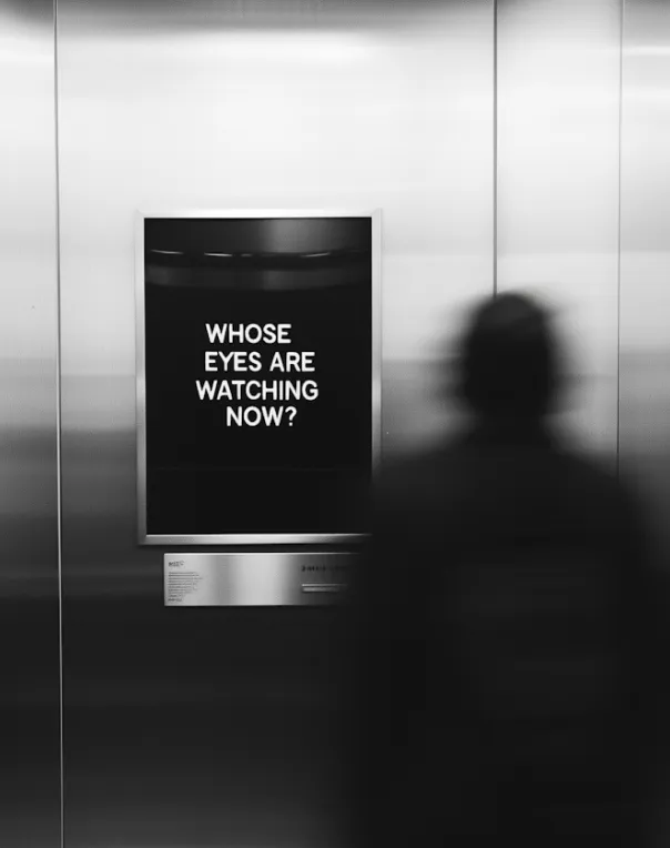 Black and white photo of a person standing in front of a metallic wall with a framed sign that reads 'WHOSE EYES ARE WATCHING NOW?'