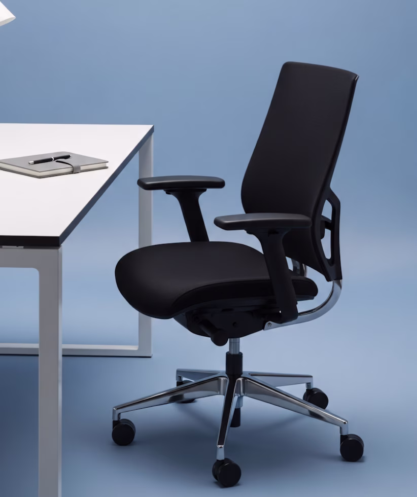 Modern black ergonomic office chair with armrests next to a white desk with a notebook and pen on it, set against a blue background.