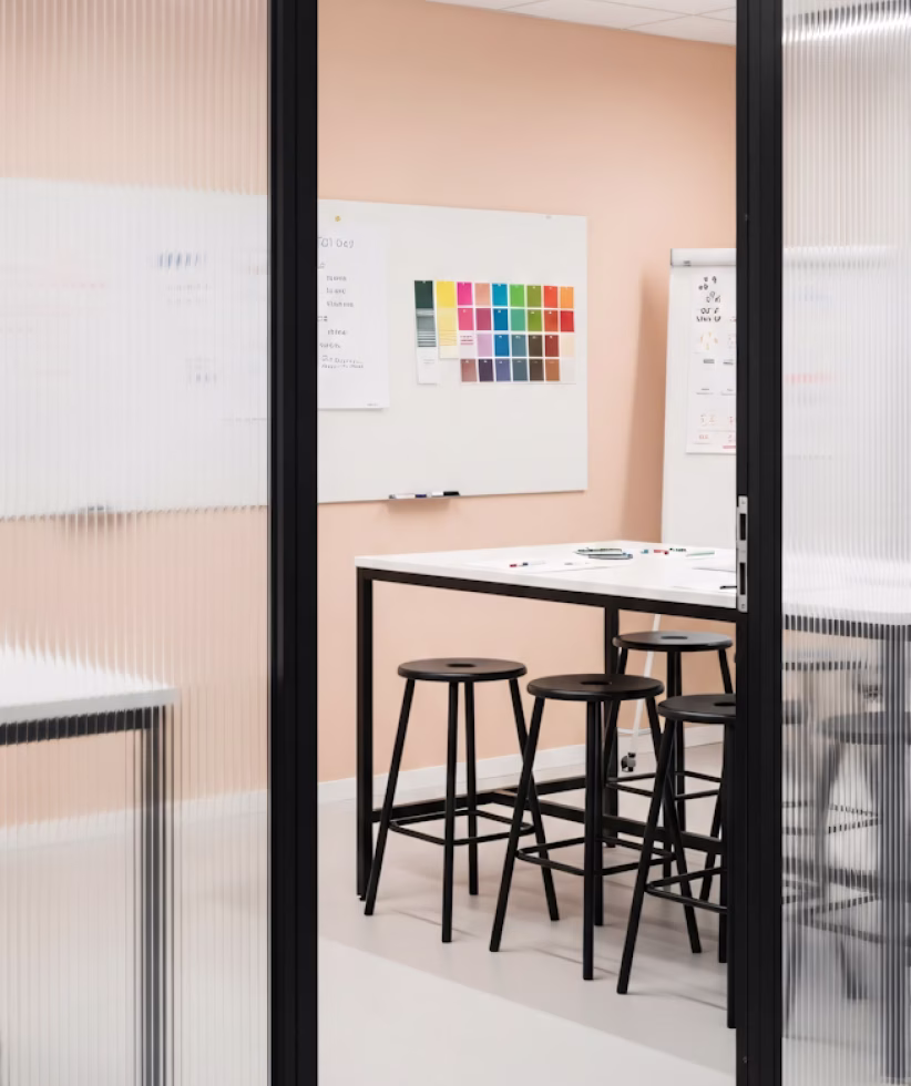 Modern office meeting room with a high white table, four black stools, and a whiteboard with color swatches on a peach wall.