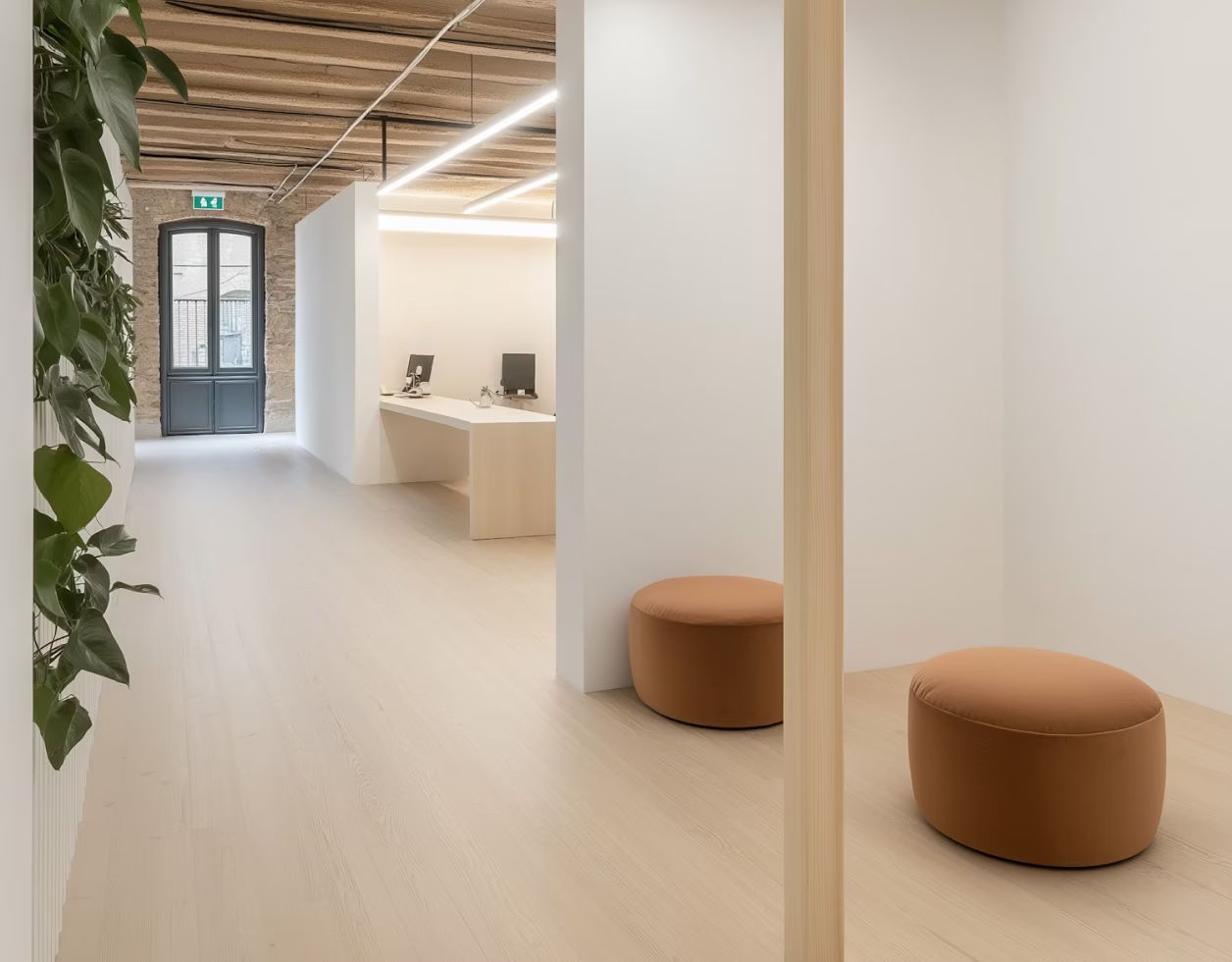 Modern, minimalist office space with light wood flooring, white walls, exposed ceiling beams, desks with computer monitors, brown round ottomans, and a large window door at the far end.
