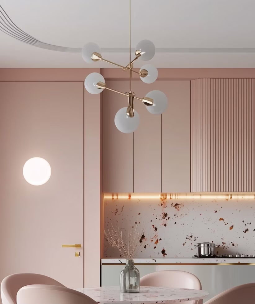 Modern kitchen with light pink cabinetry, a round marble table with pink chairs, a glass vase with dried branches, and a gold chandelier with frosted glass globes.