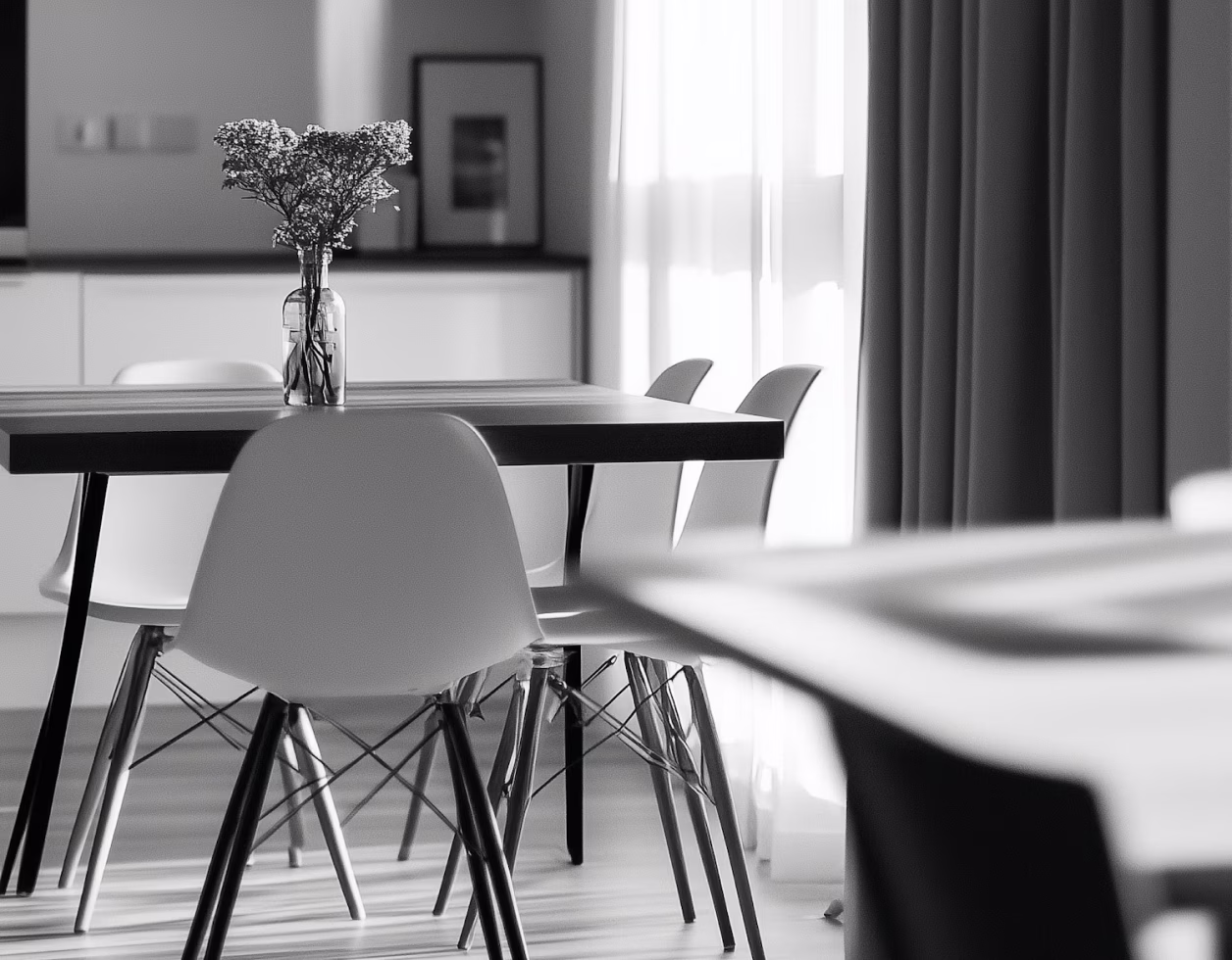 Modern dining table with white chairs and a vase of flowers in a bright room with sheer curtains.
